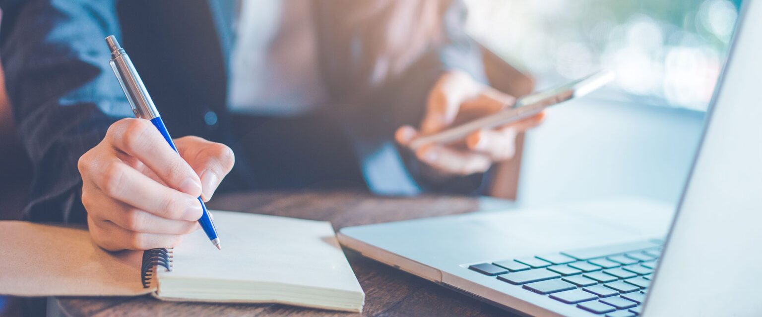 man on his phone with a laptop making notes in a notepad